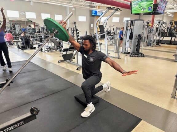 A man performs a half-kneeling landmine press with a weight plate in a gym, balancing on one knee and extending his opposite arm for stability.