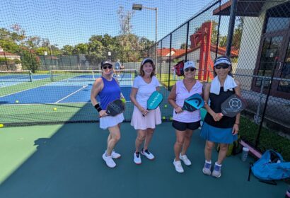 Four women stand on an outdoor pickleball court, each holding a paddle, posing for a photo on a sunny day.