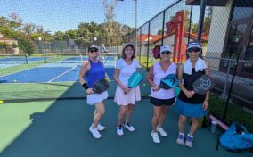 Four women in sports attire stand on a pickleball court holding paddles, with a net and another pair playing in the background on a sunny day.