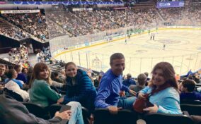 Four people sit in arena seats, smiling at the camera, with a hockey game and a large crowd visible in the background.