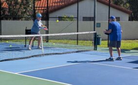 Two men play pickleball on an outdoor court, with one hitting the ball over the net while the other prepares to return the shot.