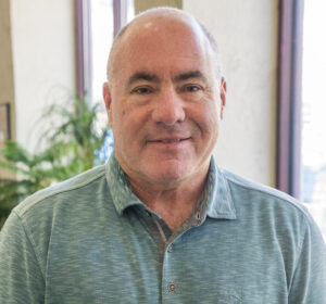 A middle-aged man with short gray hair wearing a green polo shirt stands indoors in front of large windows and greenery.