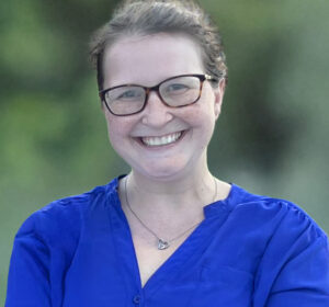 A person wearing glasses and a blue shirt is smiling at the camera, standing outdoors with a blurred green background.