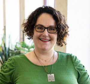 A person with short curly hair and glasses, wearing a green top and pendant necklace, stands indoors and smiles at the camera.