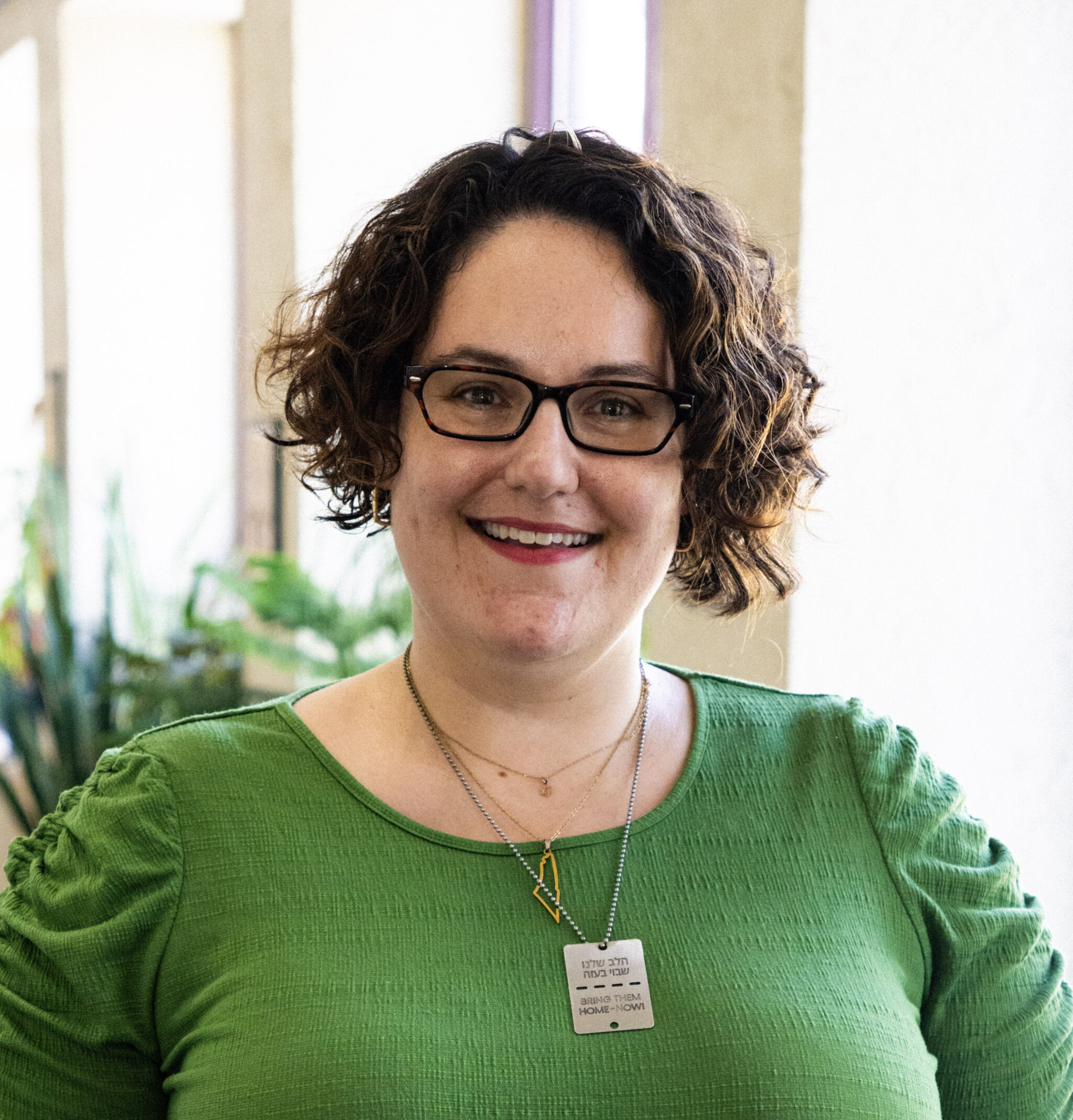 A person with short curly hair and glasses, wearing a green top and pendant necklace, stands indoors and smiles at the camera.