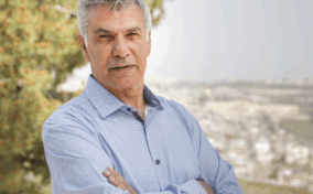 An older man with gray hair stands outdoors with arms crossed, wearing a light blue shirt. A tree and an out-of-focus cityscape appear in the background.