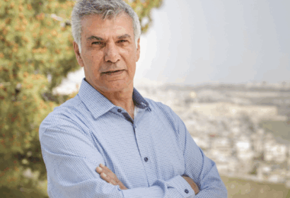 An older man with gray hair stands outdoors with arms crossed, wearing a light blue shirt. A tree and an out-of-focus cityscape appear in the background.