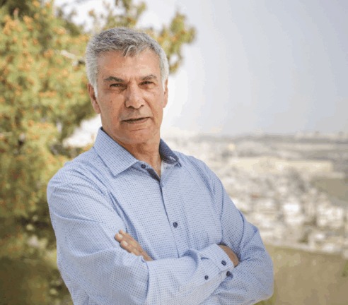 An older man with gray hair stands outdoors with arms crossed, wearing a light blue shirt. A tree and an out-of-focus cityscape appear in the background.