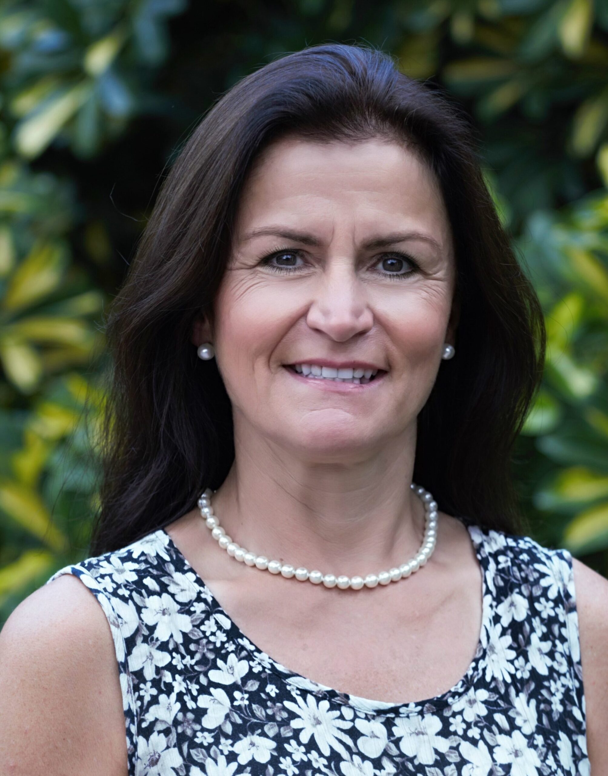Woman with long dark hair wearing a black and white floral dress and a pearl necklace stands in front of leafy green foliage, smiling at the camera.