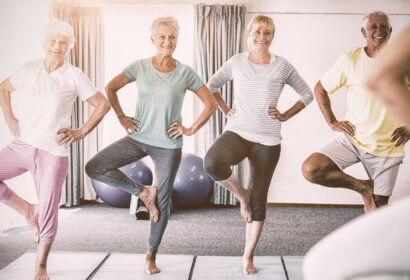 Four older adults in workout clothes stand on yoga mats, balancing on one leg in a bright exercise room with large windows and fitness balls in the background.