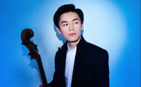 A man in formal attire holds a cello and looks to the side against a blue background.