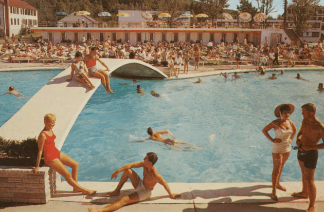 People relax, swim, and sunbathe around a crowded outdoor pool at a resort; lounge chairs, umbrellas, and a bridge are visible.