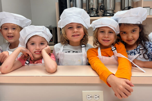 Five young children wearing chef hats and aprons stand together behind a counter, smiling at the camera in a kitchen setting.