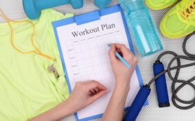 Person writing on a workout plan clipboard, surrounded by exercise clothes, a whistle, dumbbell, water bottle, shoes, and a jump rope.