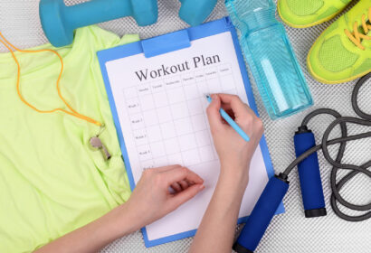 Person writing on a workout plan clipboard, surrounded by exercise clothes, a whistle, dumbbell, water bottle, shoes, and a jump rope.