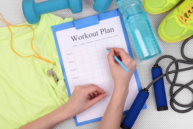 Person writing on a workout plan clipboard, surrounded by exercise clothes, a whistle, dumbbell, water bottle, shoes, and a jump rope.