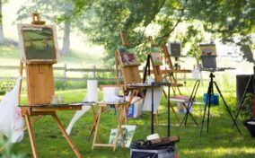 Several easels with landscape paintings set up outdoors on grass, surrounded by art supplies, under leafy trees in a sunlit area.