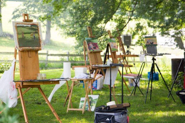 Several easels with landscape paintings set up outdoors on grass, surrounded by art supplies, under leafy trees in a sunlit area.
