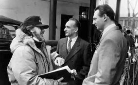 Three men stand outdoors in conversation; one man holds a folder and wears a cap and jacket, while the other two are dressed in suits.