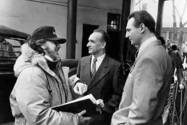 Three men stand outdoors in conversation; one man holds a folder and wears a cap and jacket, while the other two are dressed in suits.