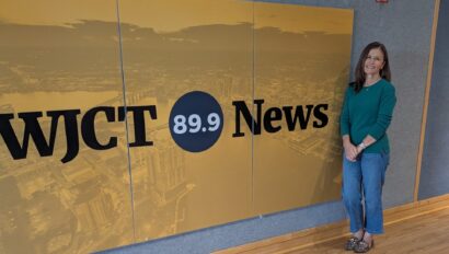 A woman stands next to a large yellow and black WJCT 89.9 News sign inside a building with blue walls and wood flooring.