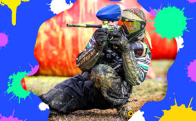 A paintball player in full gear aims a paintball gun while kneeling on the ground, with colorful paint splatters in the background.