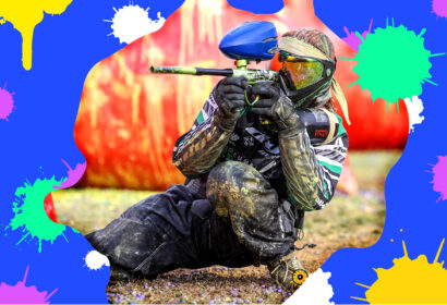 A paintball player in full gear aims a paintball gun while kneeling on the ground, with colorful paint splatters in the background.