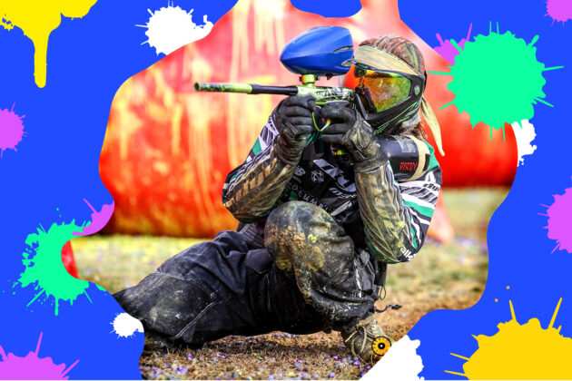 A paintball player in full gear aims a paintball gun while kneeling on the ground, with colorful paint splatters in the background.