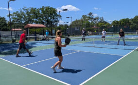 Four people play pickleball on outdoor courts under a clear sky, with two on each side of the net and others visible in the background.