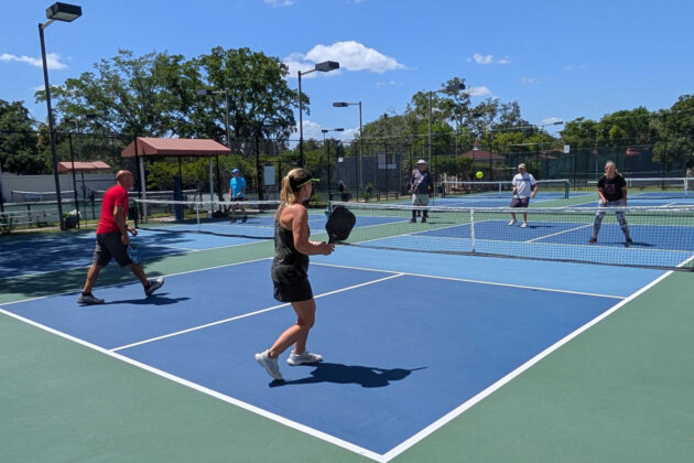 Four people play pickleball on outdoor courts under a clear sky, with two on each side of the net and others visible in the background.