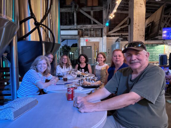 Seven people sit around a table with food and drinks in a brewery or industrial-style venue, smiling at the camera. Brewing tanks and menu boards are visible in the background.