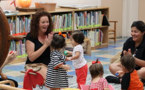 A group of adults and toddlers interact and play together in a brightly lit room with bookshelves and toys.