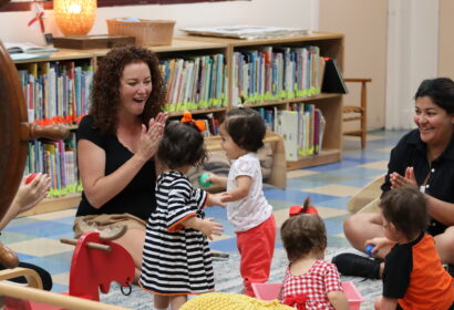 A group of adults and toddlers interact and play together in a brightly lit room with bookshelves and toys.
