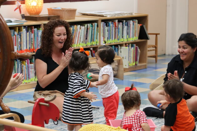 A group of adults and toddlers interact and play together in a brightly lit room with bookshelves and toys.
