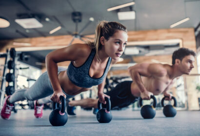Two people exercising in a gym, performing push-ups while gripping kettlebells on the floor.