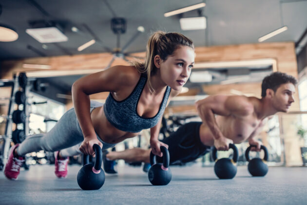 Two people exercising in a gym, performing push-ups while gripping kettlebells on the floor.