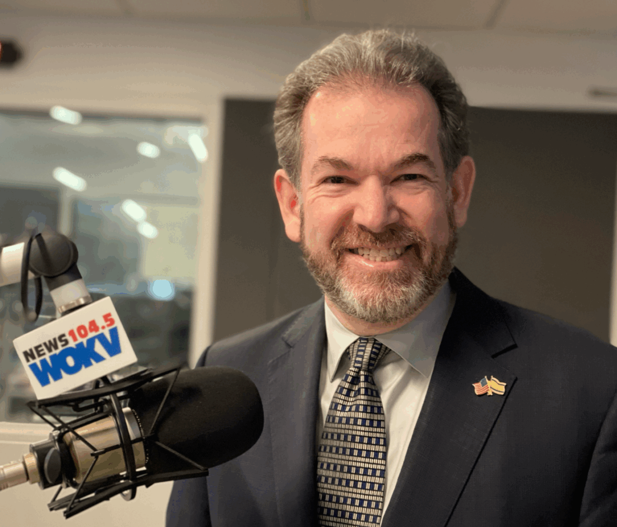 A man in a suit and tie stands next to a microphone labeled "NEWS 104.5 WOKV" in a radio studio.
