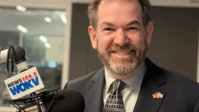 A man in a suit and tie stands next to a microphone labeled "NEWS 104.5 WOKV" in a radio studio.