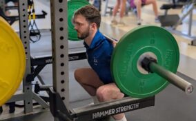 A man performs a barbell back squat with green weight plates in a gym, while other people work out in the background.