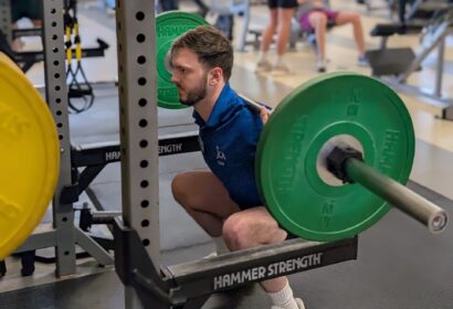 A man performs a barbell back squat with green weight plates in a gym, while other people work out in the background.