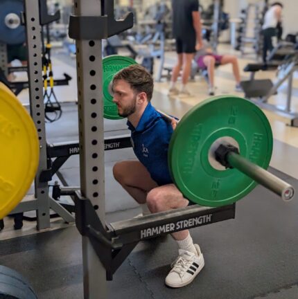 A man performs a barbell back squat with green weight plates in a gym, while other people work out in the background.