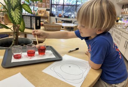 A young child uses a dropper to transfer red liquid between glasses on a tray, with a drawing and paper nearby on a classroom table.