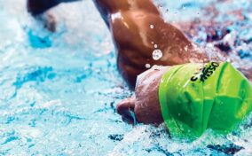 A swimmer wearing a bright green cap swims freestyle in a pool, with water splashing around their arm and head.