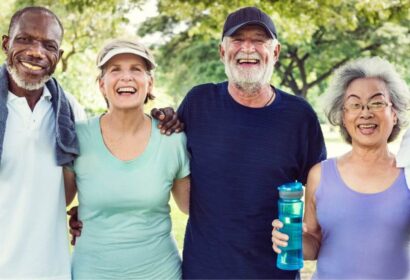 Four smiling older adults, two men and two women, stand outdoors with arms around each other, one woman holding a water bottle. Trees and greenery in the background.