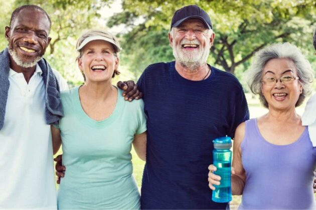 Four smiling older adults, two men and two women, stand outdoors with arms around each other, one woman holding a water bottle. Trees and greenery in the background.