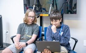 Two boys sit next to each other; one is holding and looking at a laptop while the other observes. They appear to be focused on the screen in a classroom or tech lab setting.