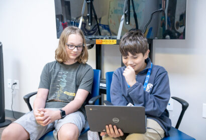 Two boys sit next to each other; one is holding and looking at a laptop while the other observes. They appear to be focused on the screen in a classroom or tech lab setting.