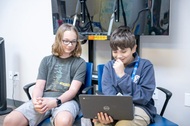 Two boys sit next to each other; one is holding and looking at a laptop while the other observes. They appear to be focused on the screen in a classroom or tech lab setting.