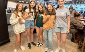 Five girls stand and smile inside a coffee shop, holding drinks. Other people are seated at tables in the background.
