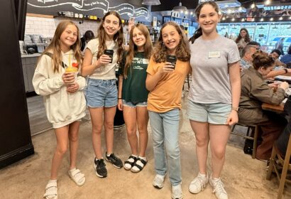 Five girls stand and smile inside a coffee shop, holding drinks. Other people are seated at tables in the background.